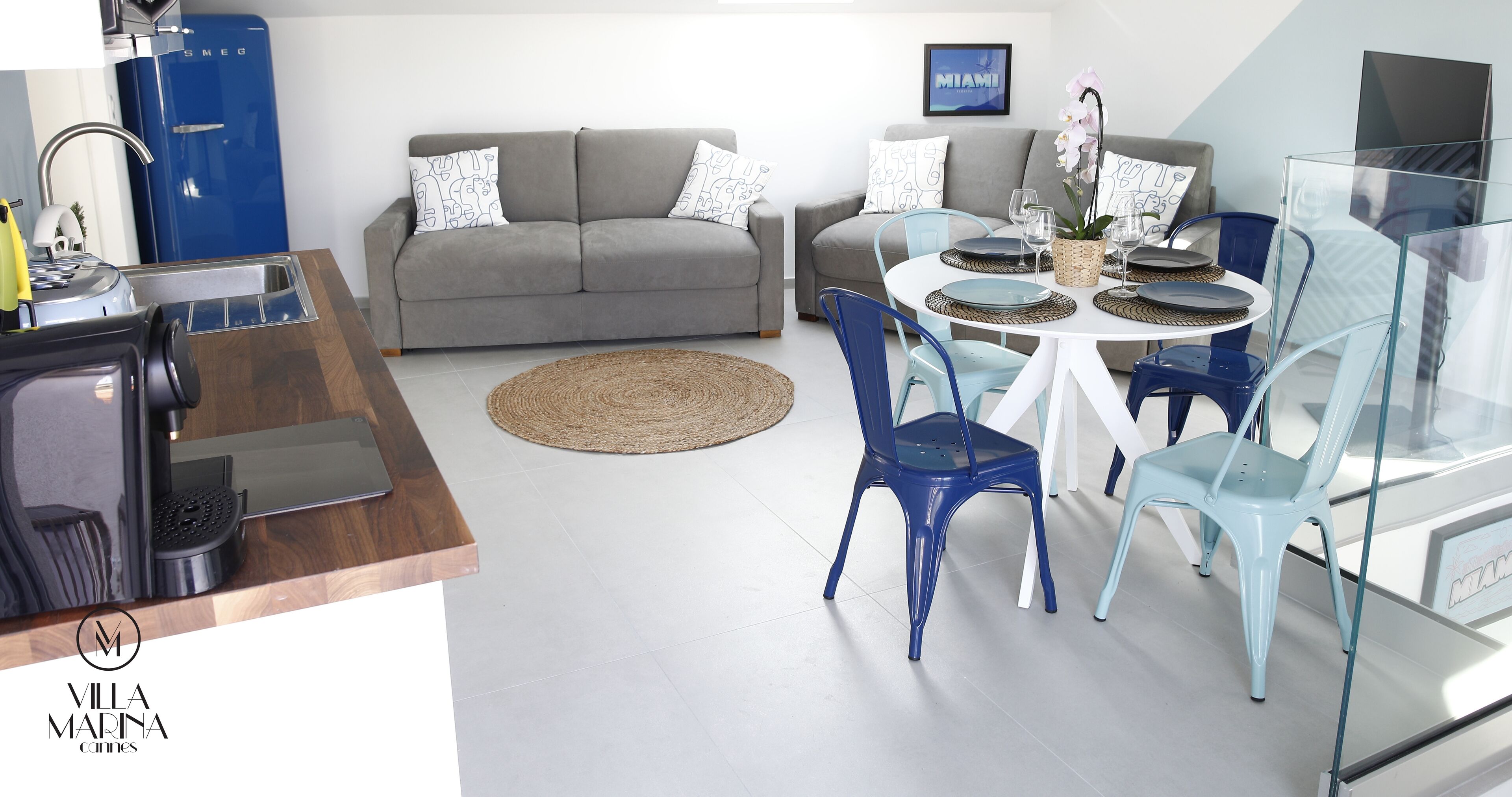 Een moderne woonkamer met een grijze bank, blauwe en witte stoelen, een ronde tafel met een geweven placemat en een blauwe waterdispenser.