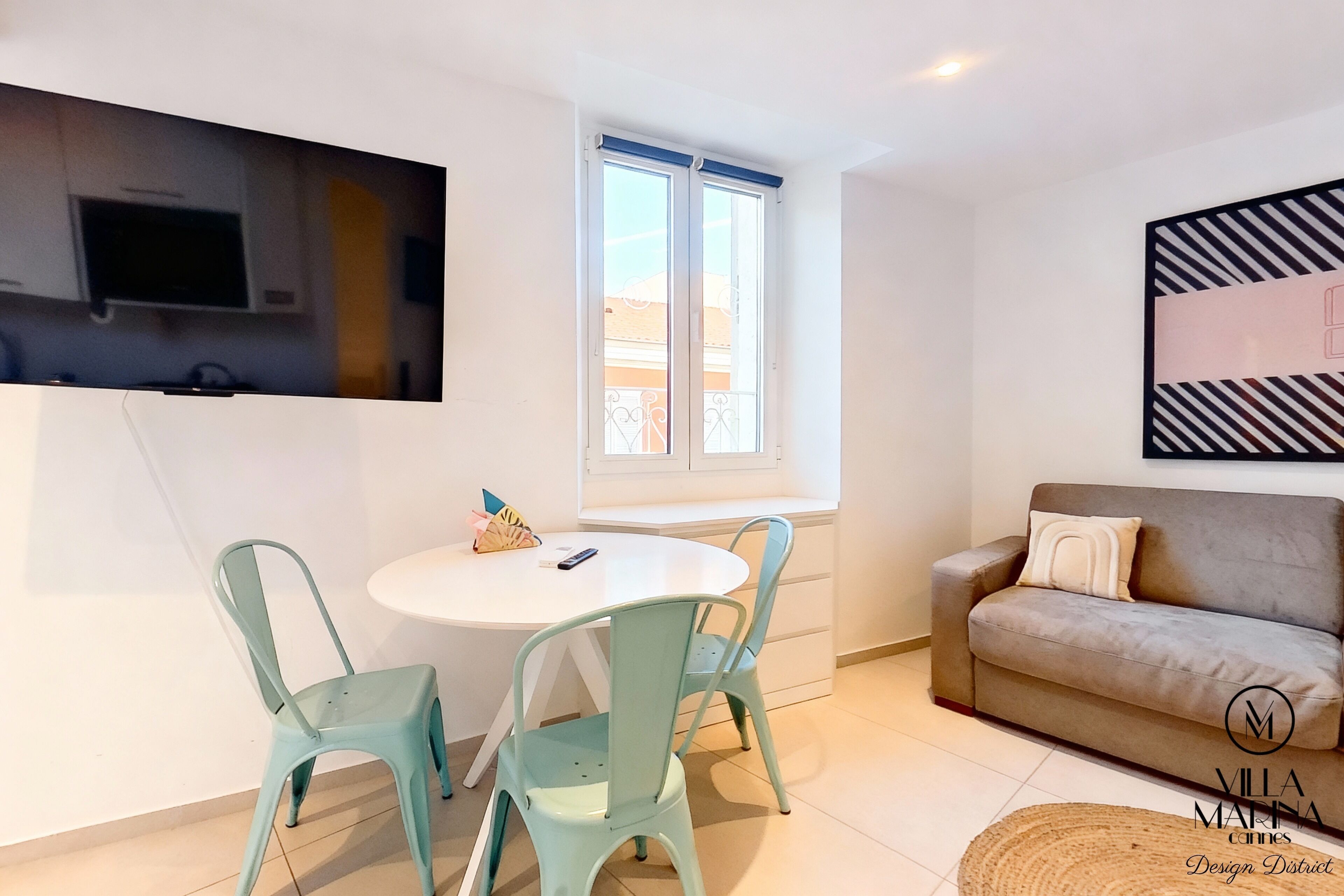 A modern living room with a round white table, two teal chairs, a grey sofa, a TV mounted on the wall, and a window with a view.