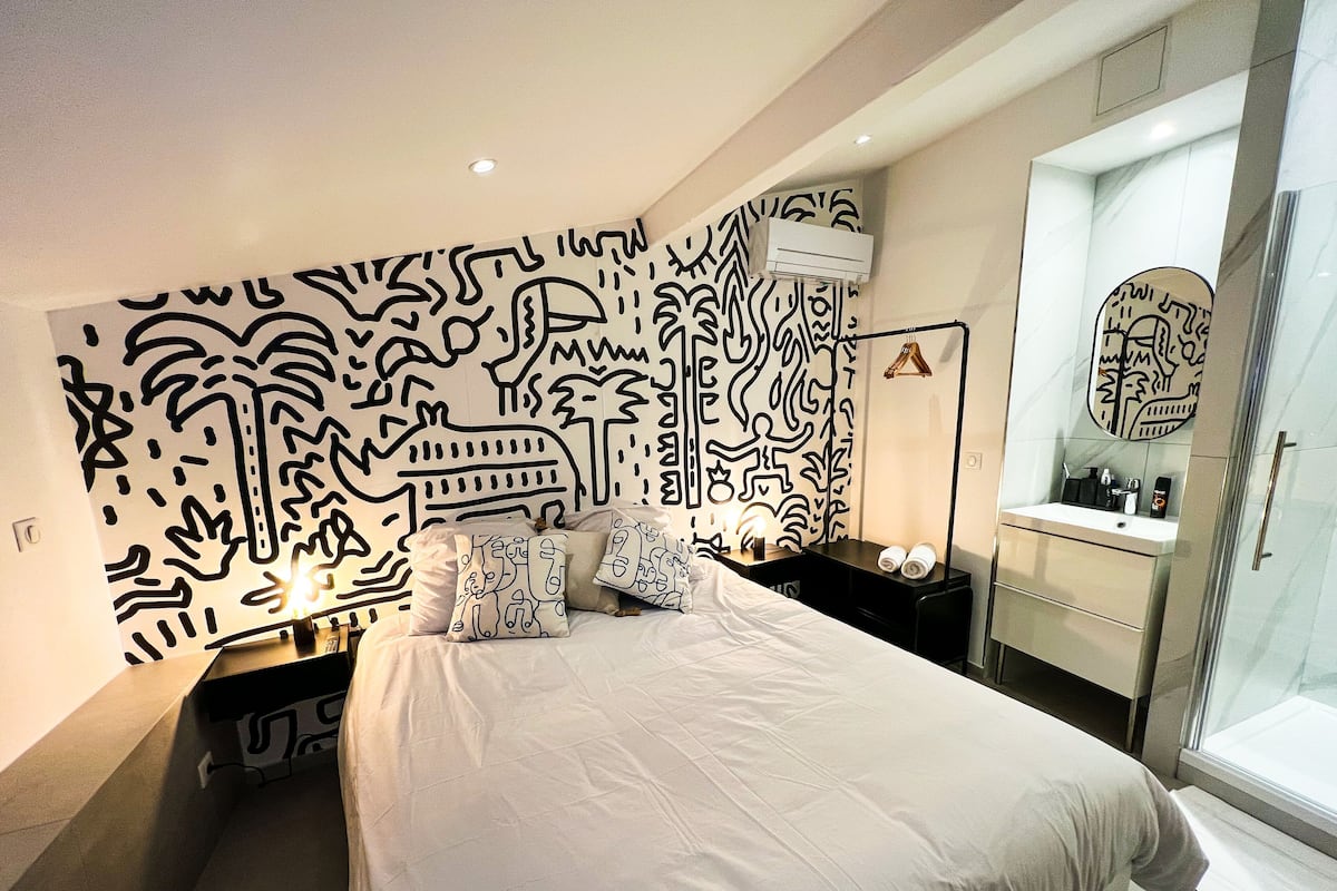 A modern bedroom with a large bed, a bedside table, a mirror, and a wall with abstract black and white patterns.