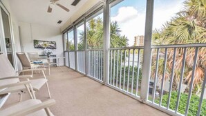 A balcony with white chairs, a ceiling fan, and a view of palm trees and buildings. - Large & Comfy Beach Apartment with Screened Patio (West Palm Beach)