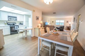 A modern kitchen with white cabinets, a microwave, and a stove. There is a dining area with a table and chairs, and a living room with a sofa and a TV.