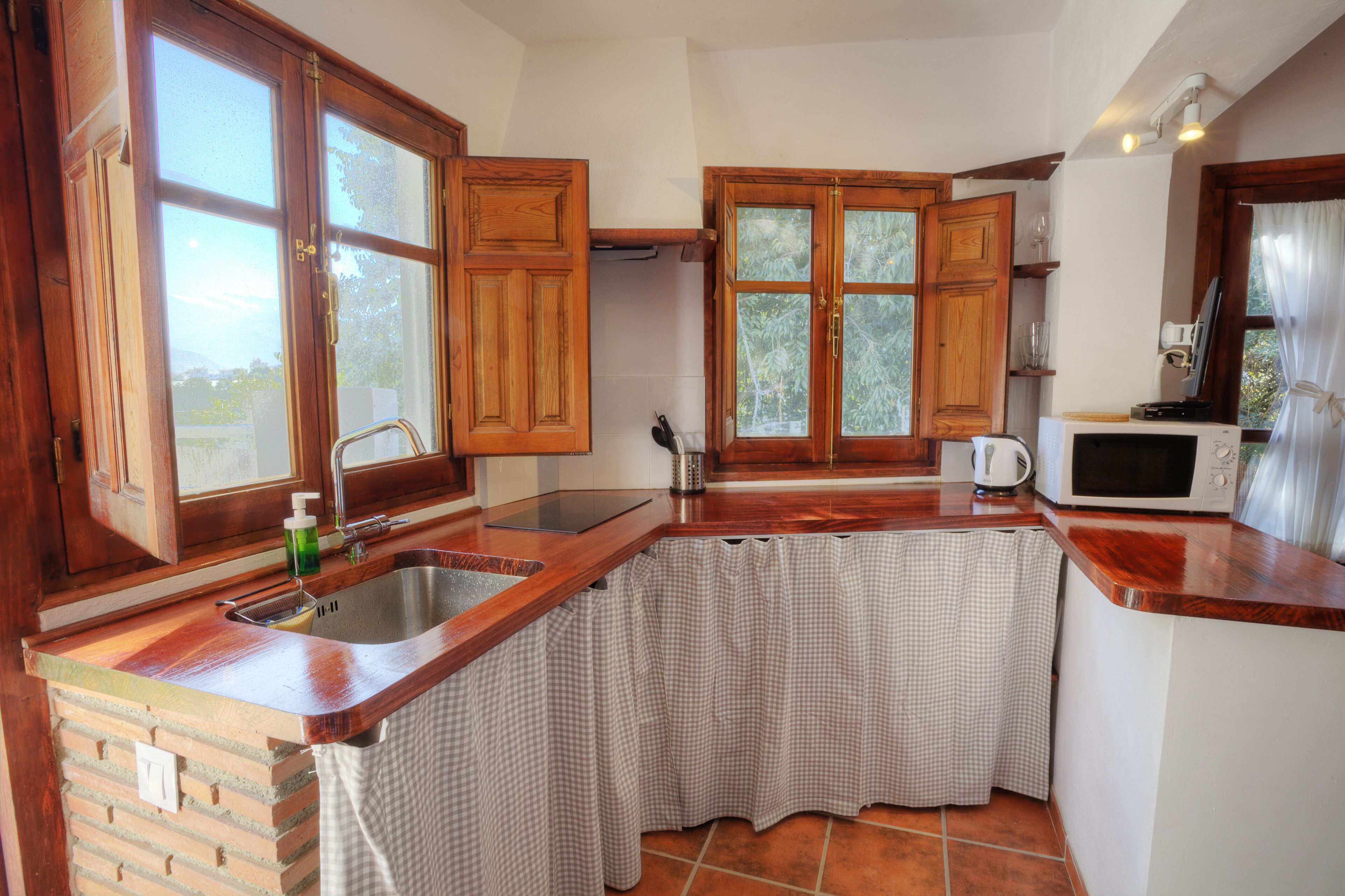Una cocina con armarios de madera, un lavamanos, un microondas y un ventanal con cortinas.