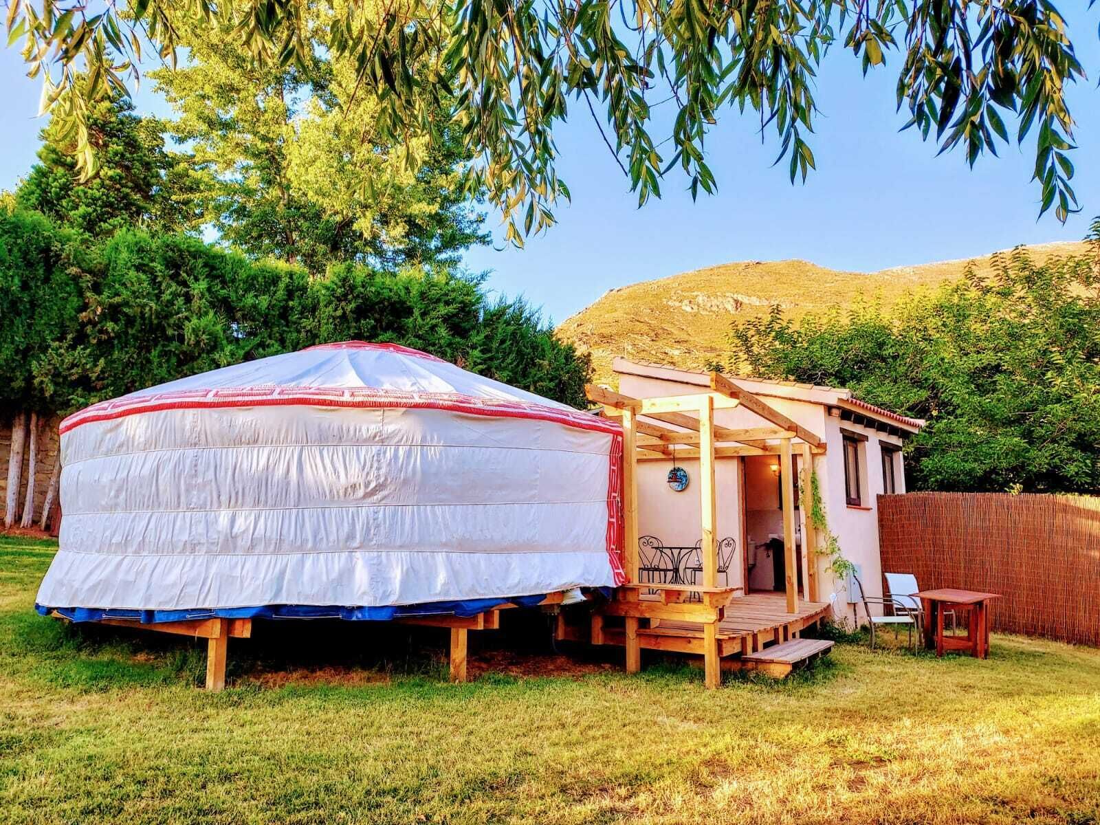A yurt with a wooden porch and a small attached structure, surrounded by trees and a grassy area.