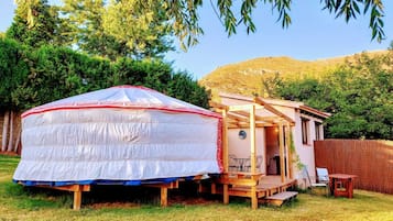 Una yurta con un porche de madera y una estructura pequeña adosada, rodeada de árboles y una zona cubierta de césped.