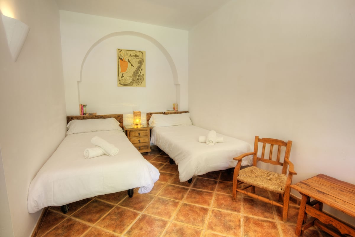 Ein Schlafzimmer mit zwei Betten, einem Holzstuhl, einem kleinen Tisch und einem Wandbild.