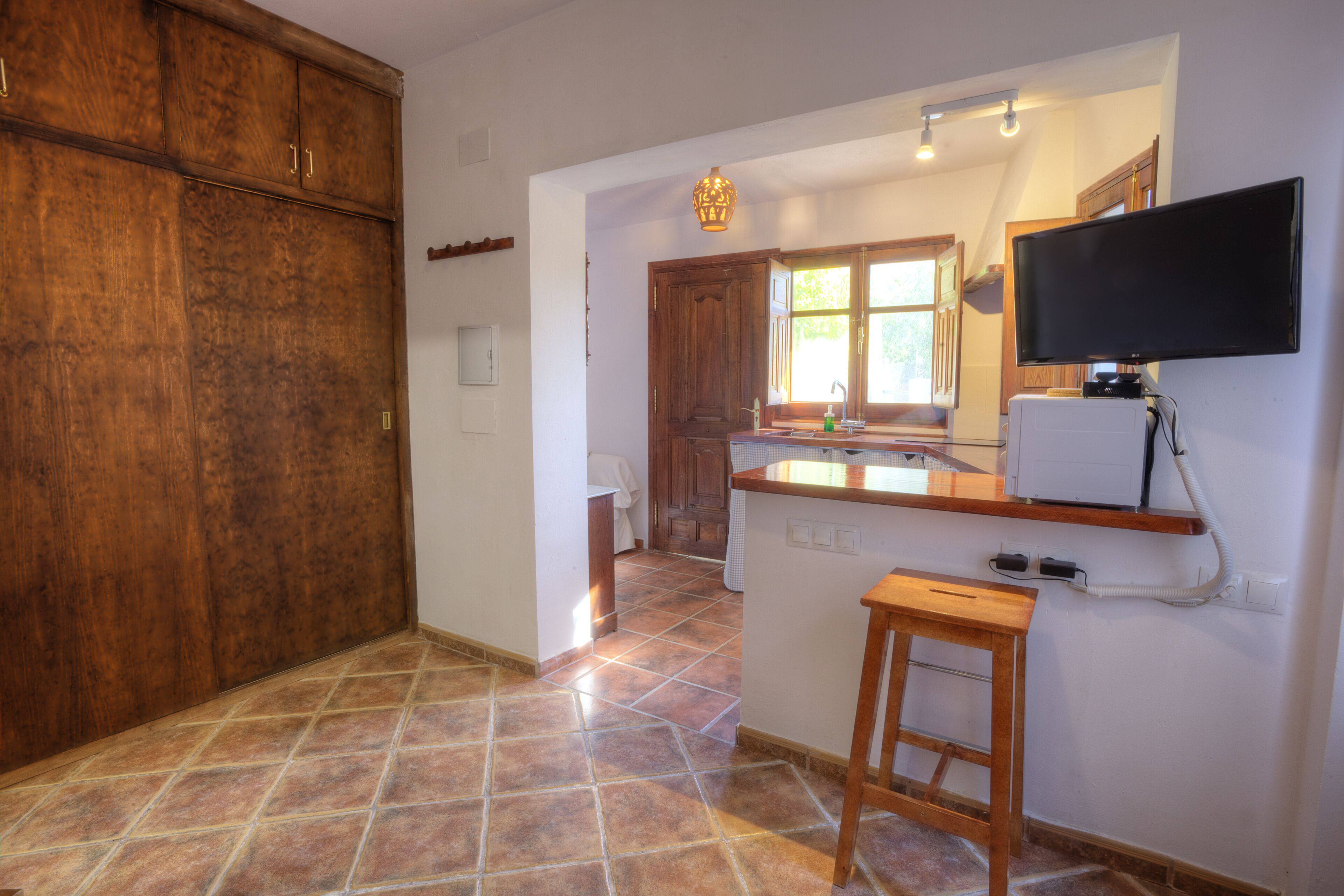 A kitchen with wooden cabinets, a tiled floor, a wooden stool, a built-in oven, and a wall-mounted television.