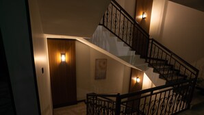 Una escalera con barandilla de hierro forjado negro, una puerta de madera al final y luces fijadas a la pared a cada lado.