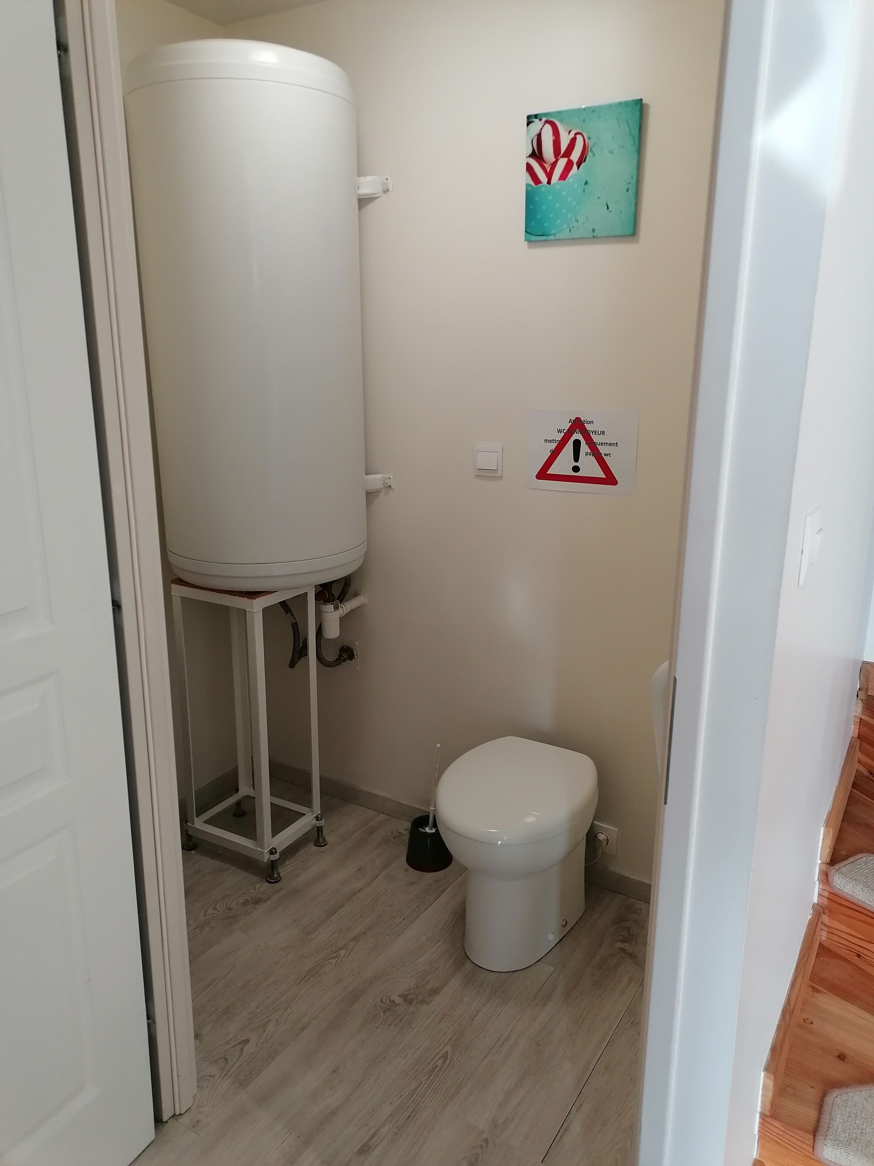 Un bagno piccolo con un water, un serbatoio d'acqua bianco, un accessorio per la pulizia a pavimento e un cartello di avvertimento sul muro.