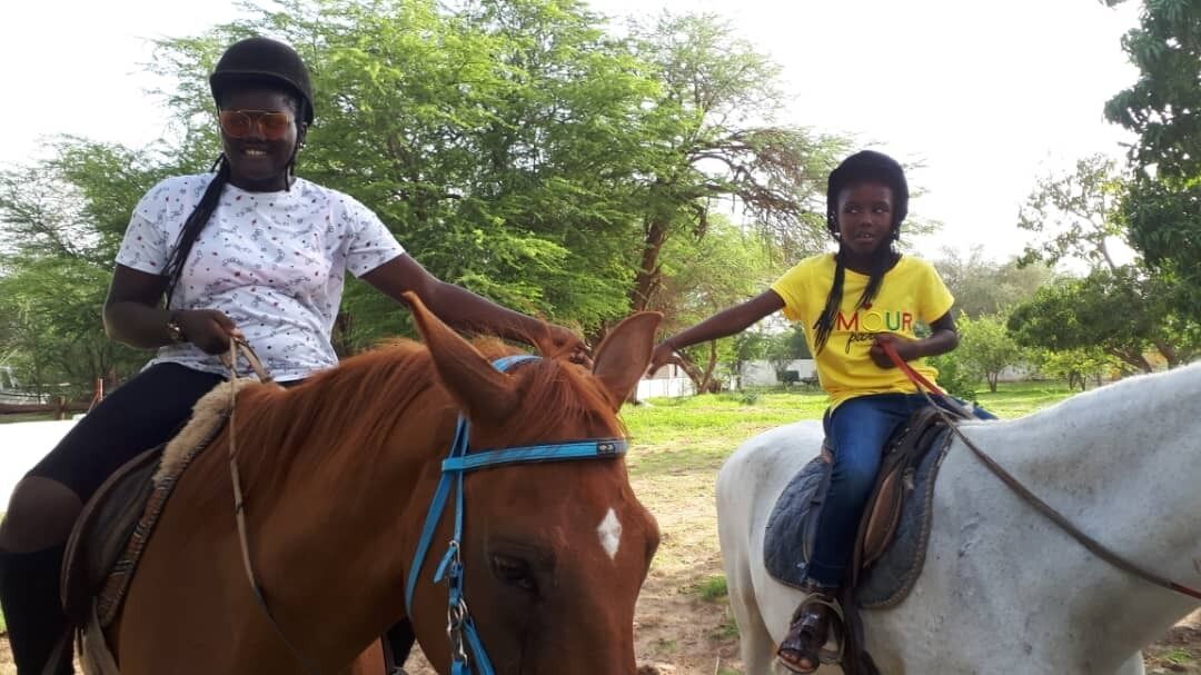 Dos personas montando a caballo en un entorno al aire libre, con árboles de fondo.