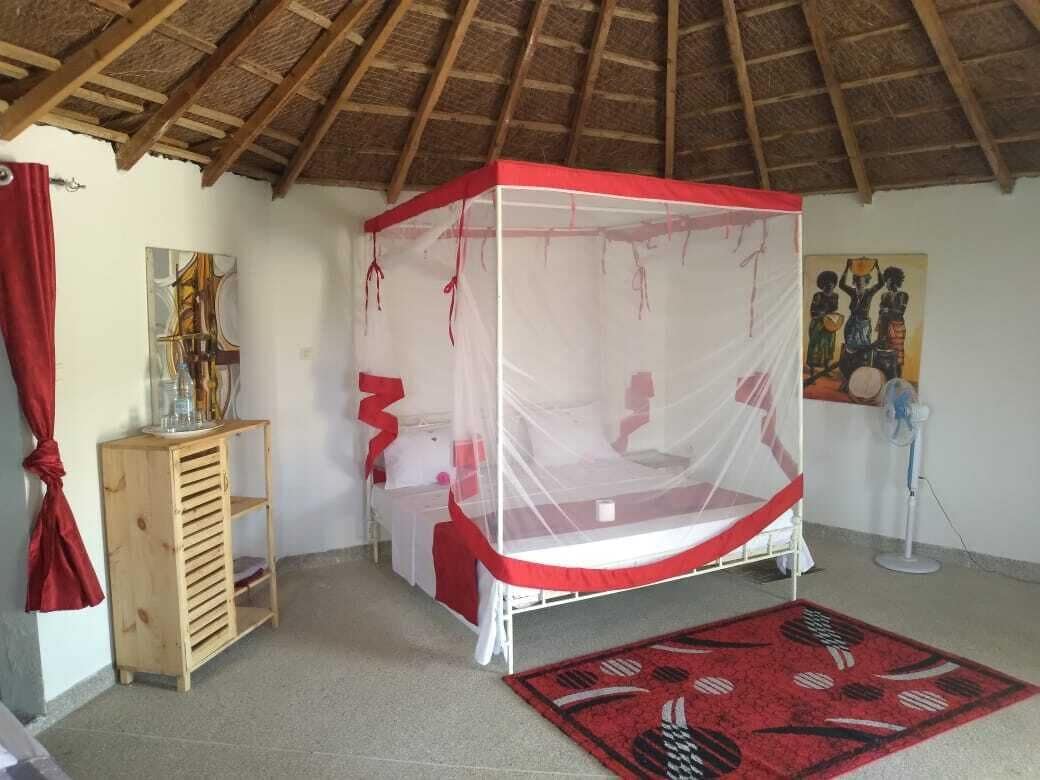 A room with a bed under a mosquito net, a wooden cabinet, a fan, and a red rug.