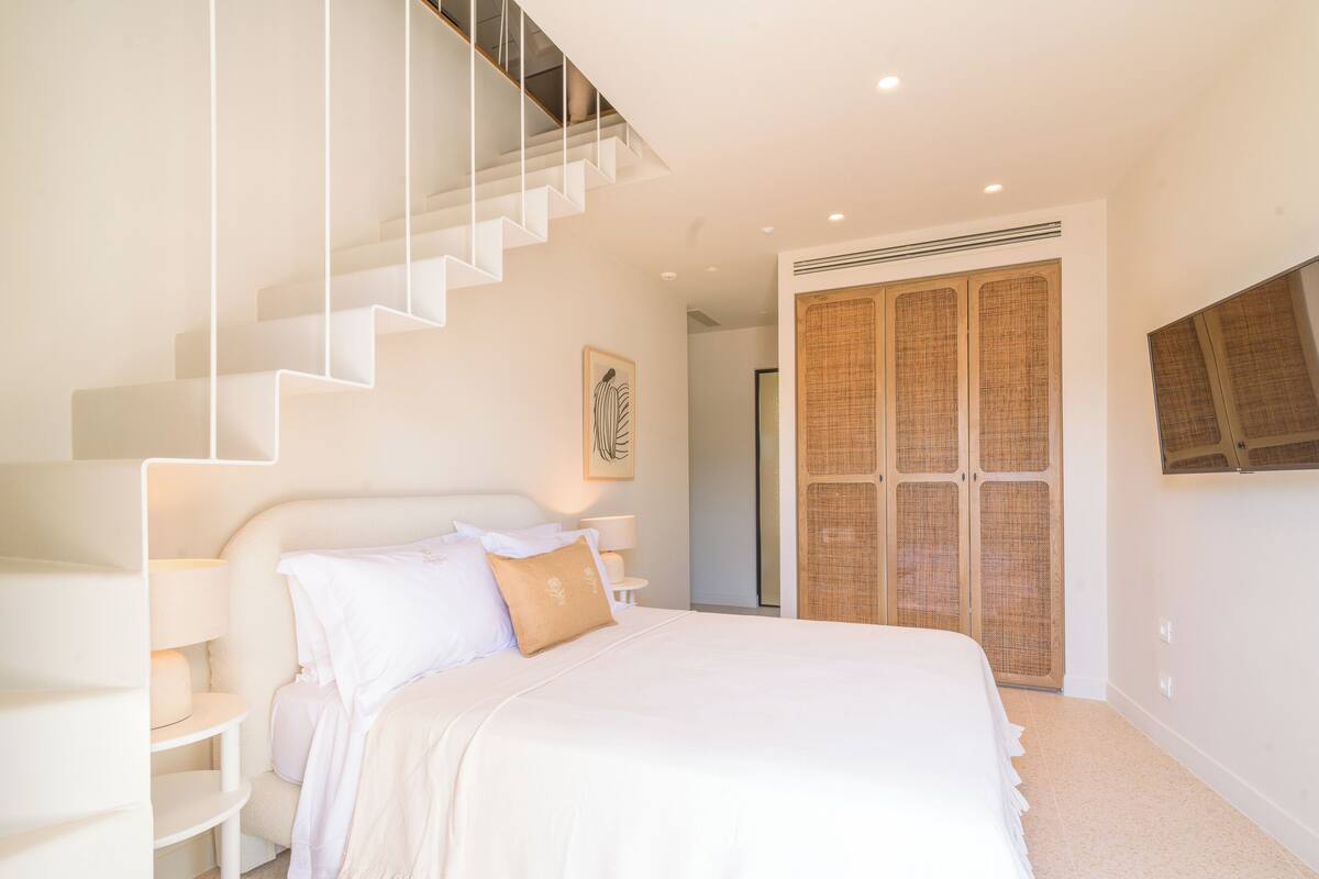 Une chambre à coucher avec un grand lit, un escalier, une table de chevet avec une lampe et une armoire dont la porte est faite d’un matériau tissé.