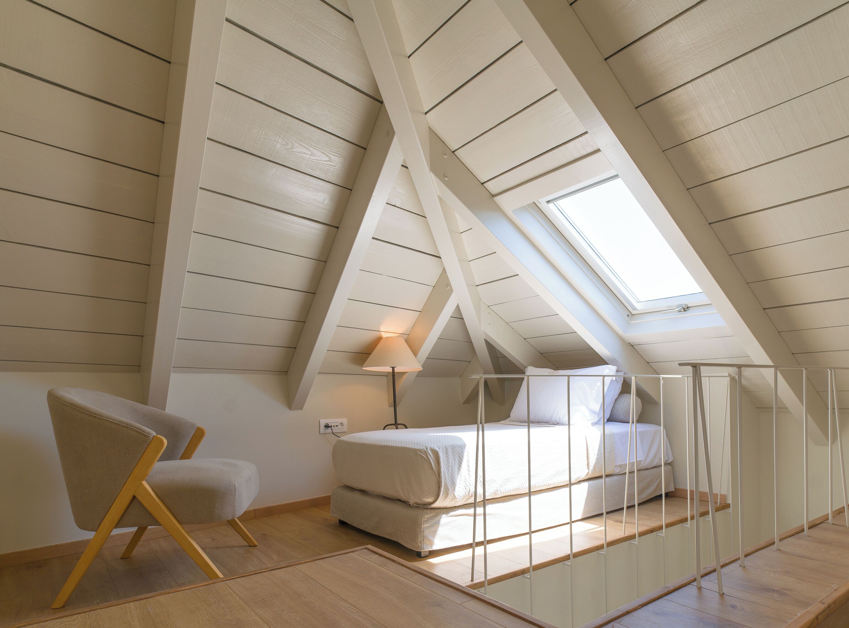 Ein Schlafzimmer mit schrägverdeckter Decke, einem Bett, einem Sessel und einem Fenster.