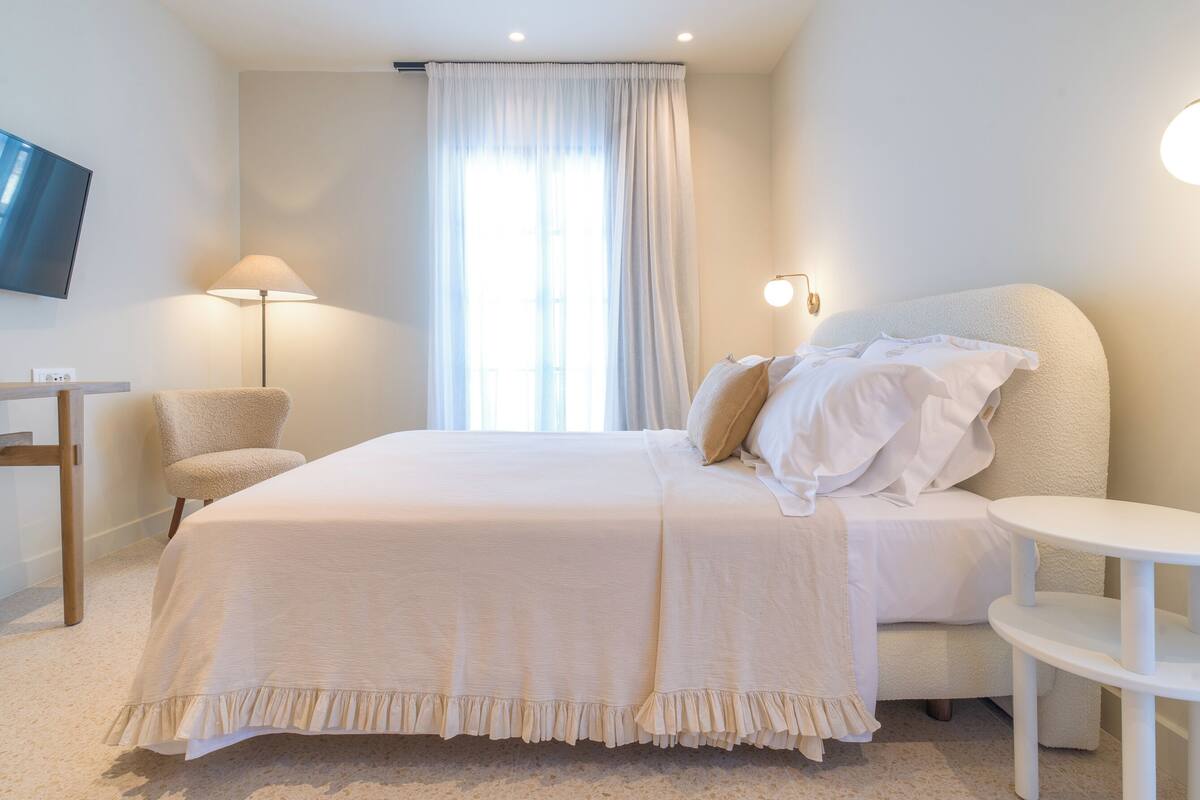 Une chambre d’hôtel avec un lit, une chaise, une petite table, un téléviseur et une fenêtre avec des rideaux.