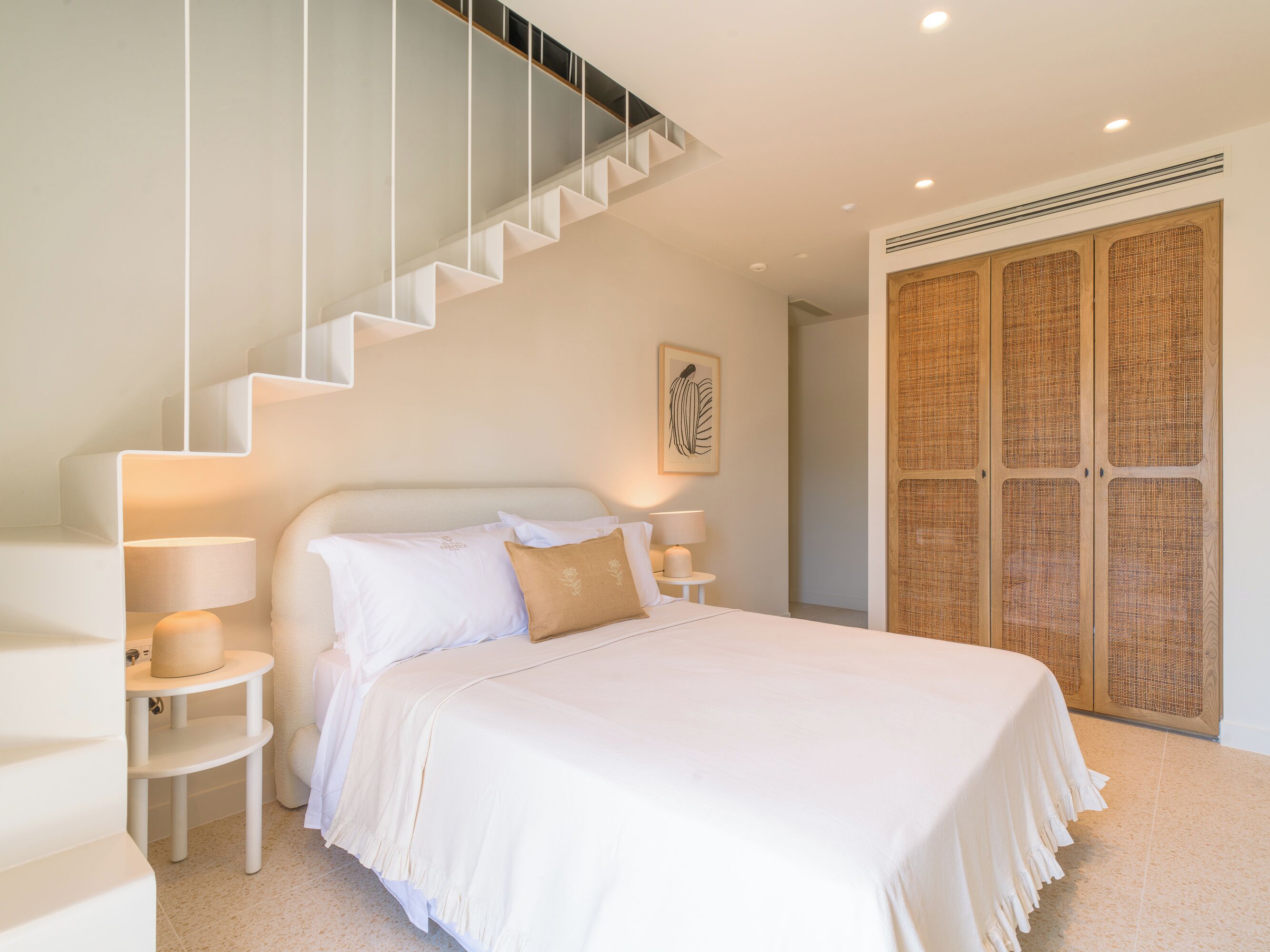 Un dormitorio con cama, mesitas de noche, lámpara, una obra de arte en la pared, una escalera y un armario de madera.
