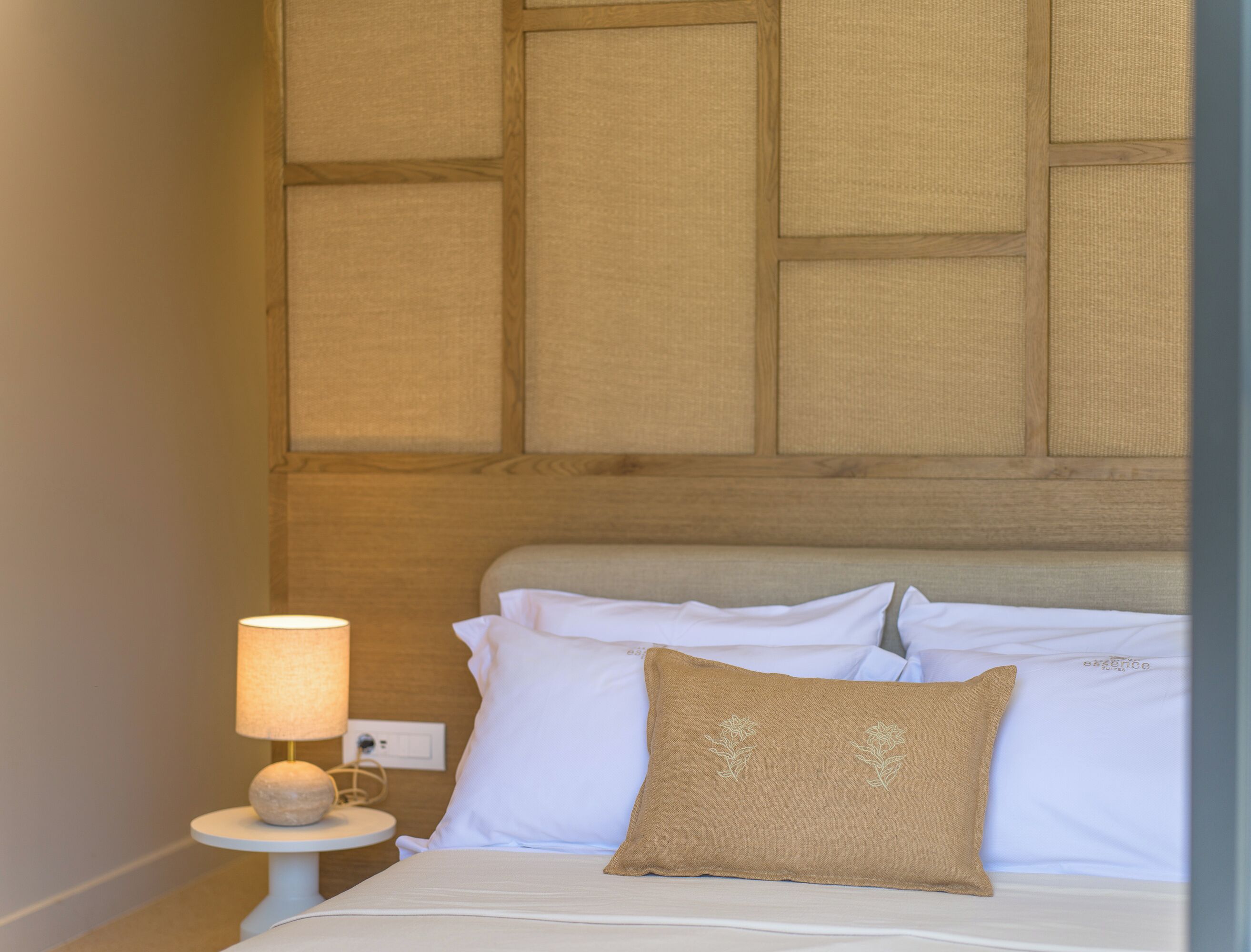 Una cama bien tendida con ropa blanca y una única almohada marrón, una mesa redonda con una lámpara, y una pared con diseño de paneles de madera.
