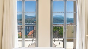 A bedroom with a large bed, two sliding glass doors leading to a balcony with a view of a lake and mountains. - Argostoli Essence Suites (Kefalonia)