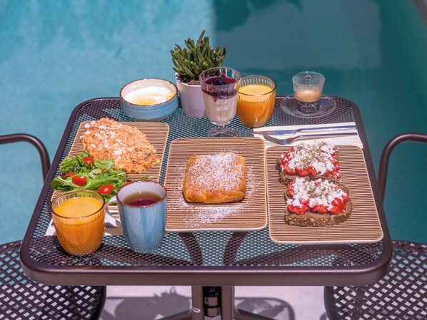 A table with a variety of food items and drinks, including a salad, a pastry, and a sandwich, set against a poolside backdrop. - Argostoli Essence Suites (Kefalonia)