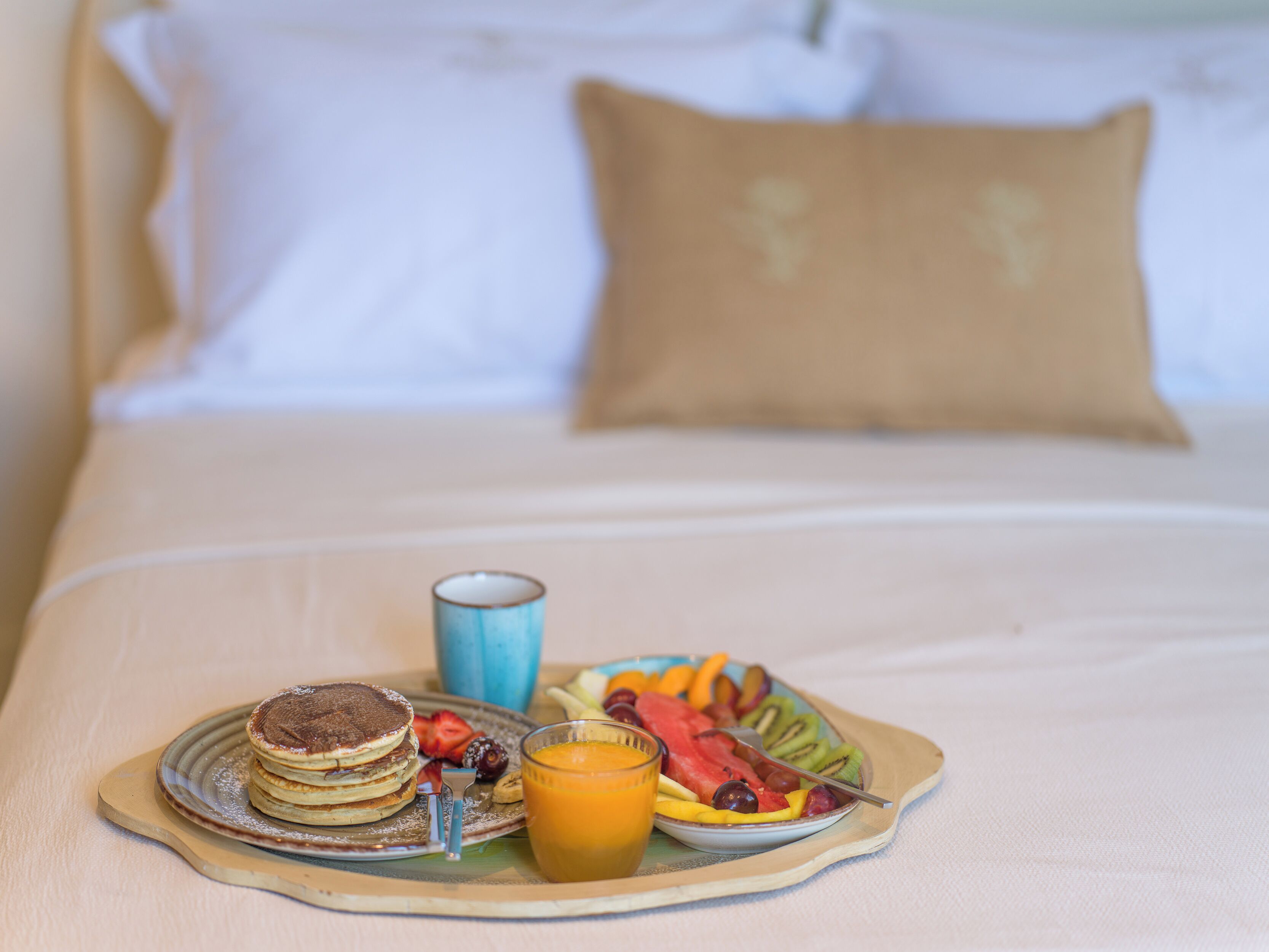 A breakfast tray with pancakes, fruit, and orange juice on a bed with white bedding and a brown pillow.