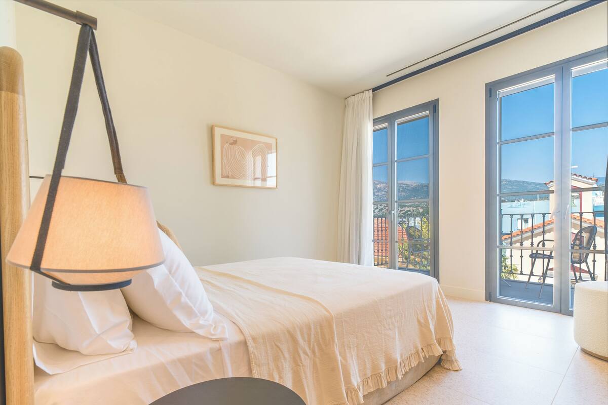 Une chambre à coucher avec un lit, une lampe suspendue, un tableau encadré au mur et un balcon donnant sur des immeubles.