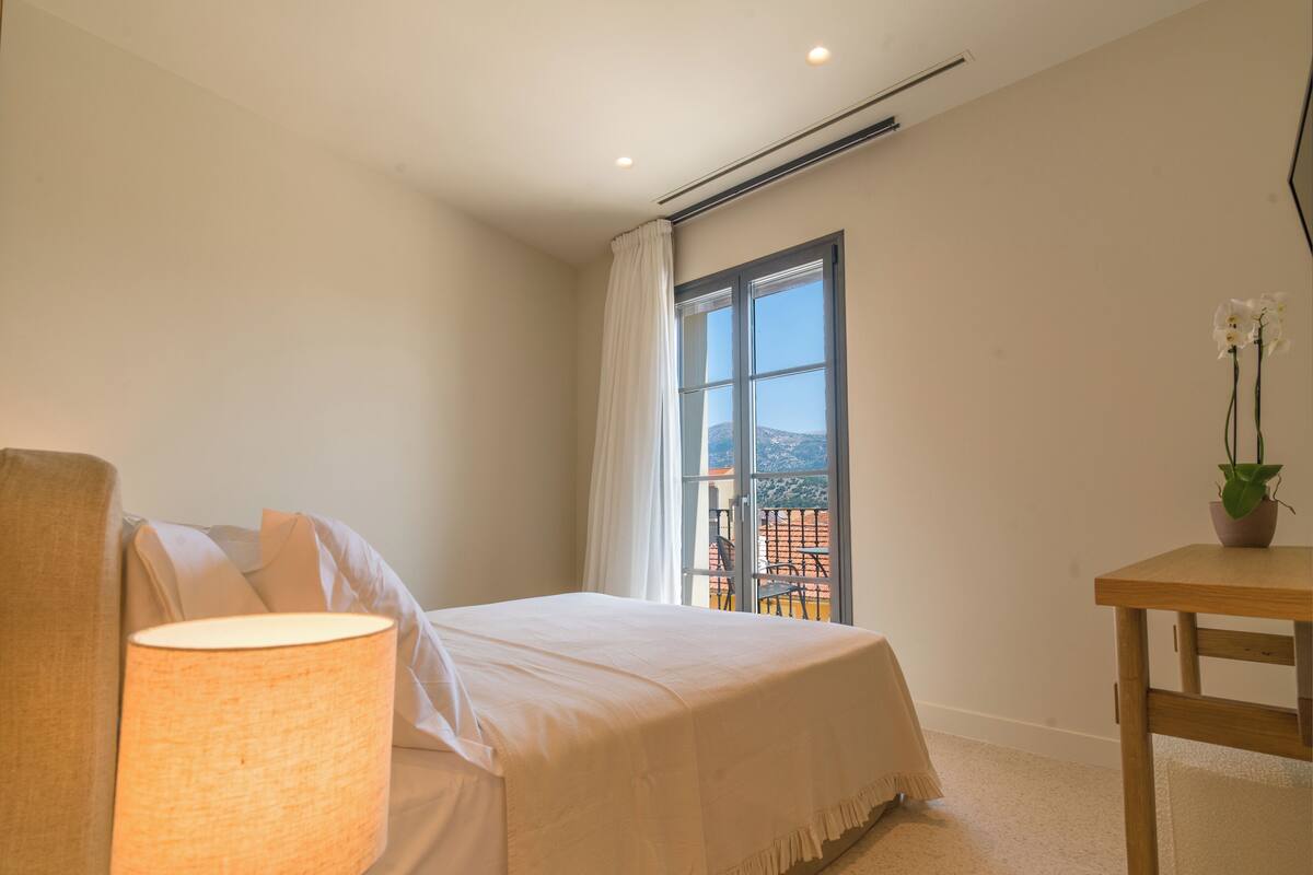 Une chambre à coucher comprenant un lit, une table de chevet, une lampe, une fenêtre avec des rideaux et une vue sur l’extérieur.