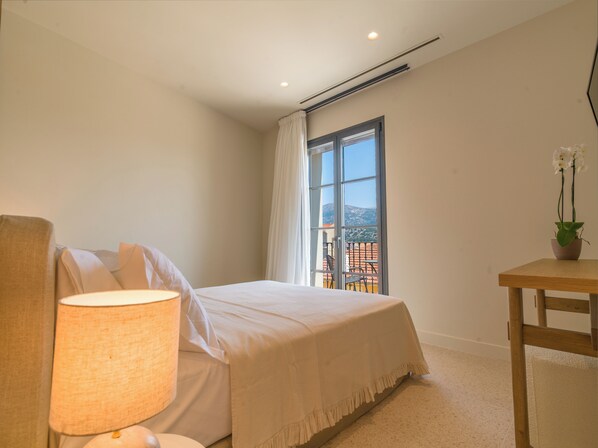 Un dormitorio con cama, mesita de noche, lámpara, ventana con cortinas y vistas al exterior.