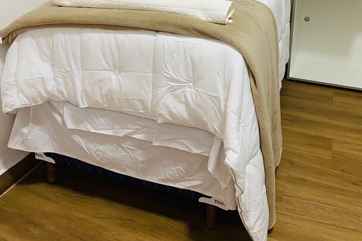 A single bed with a white duvet and a beige blanket, a white pillow, and a wooden floor.