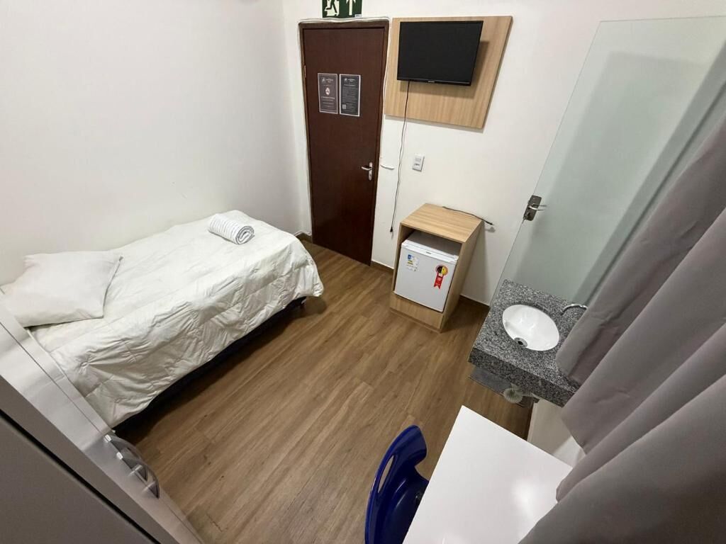 Quarto pequeno com cama, porta de madeira, televisão na parede, armário pequeno e banheiro com pia e espelho.
