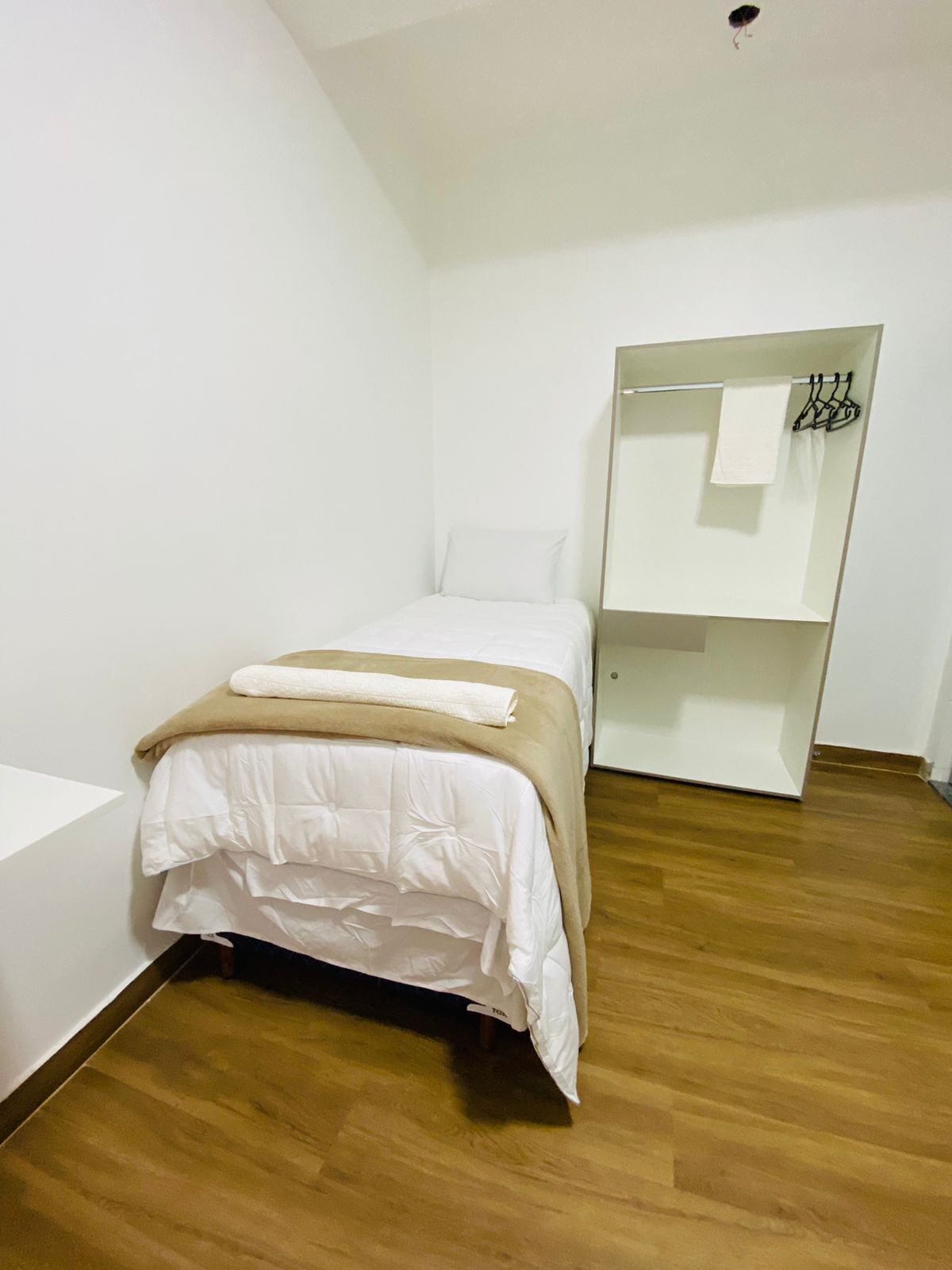 A single bed with a beige blanket and white pillow in a room with wooden flooring and white walls.