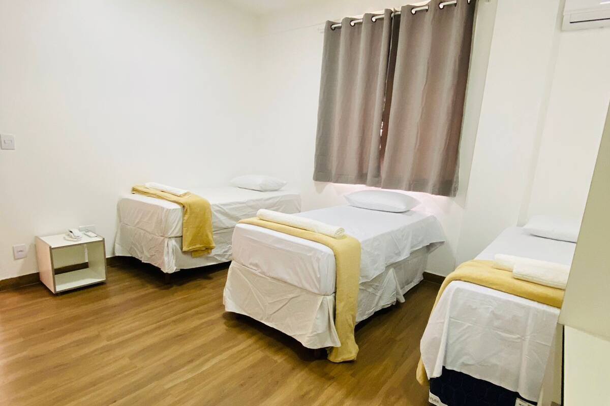 A hotel room with three single beds, a small bedside table, a nightstand, and a window with curtains.