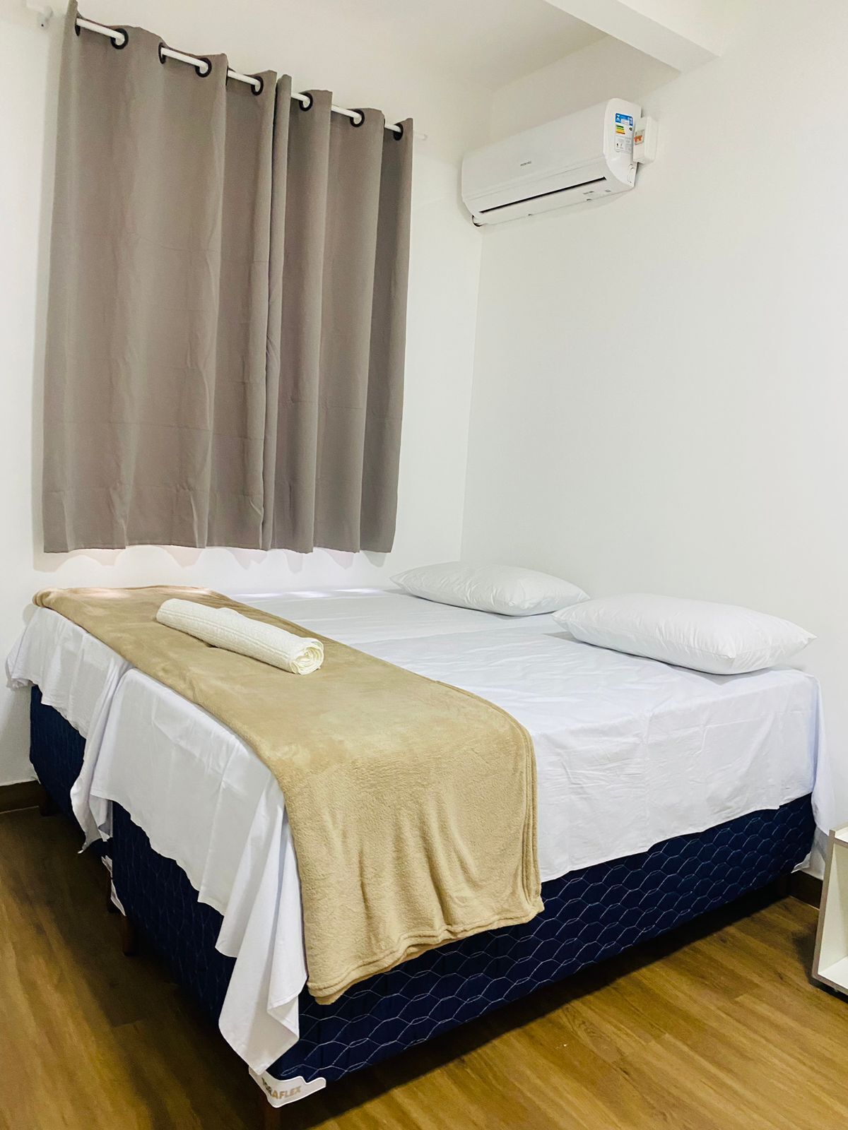 A single bed with a beige blanket, white pillows, and a blue patterned bedspread. There is a white air conditioning unit on the wall above the bed.
