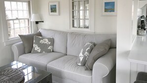 A living room with a striped sofa, a glass coffee table, and a wicker chair. There are two windows with curtains, a lamp, and framed pictures on the wall. - Barn/cottage (Nantucket)