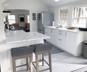 Une cuisine moderne avec des meubles blancs, un plan de travail en marbre et un réfrigérateur encastré. On y trouve deux tabourets de bar et une fenêtre qui laisse entrer la lumière naturelle.