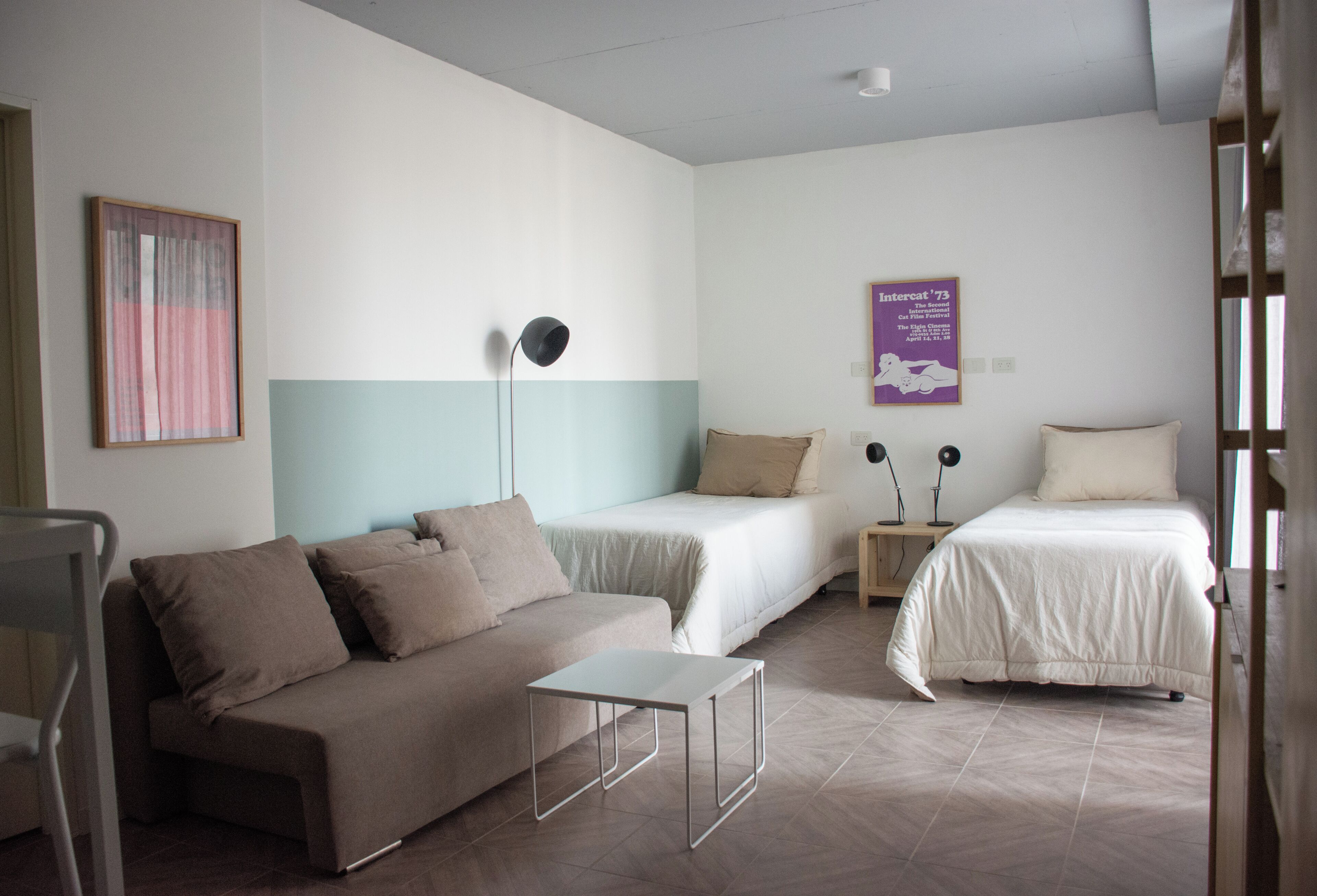 A room with a sofa, two single beds, a small table, a floor lamp, and a poster on the wall.
