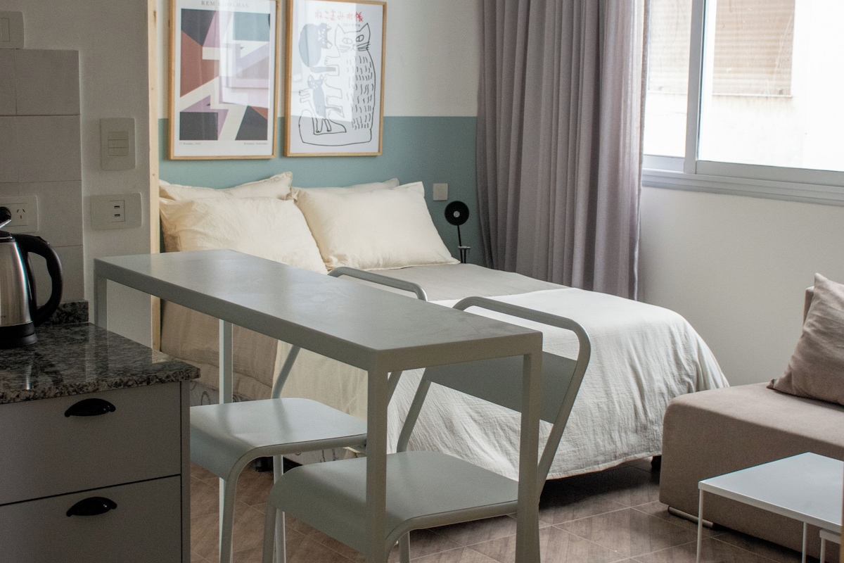 A modern, well-lit bedroom with a bed, a desk, and a chair. There is a window with curtains, a radiator, and artwork on the wall.