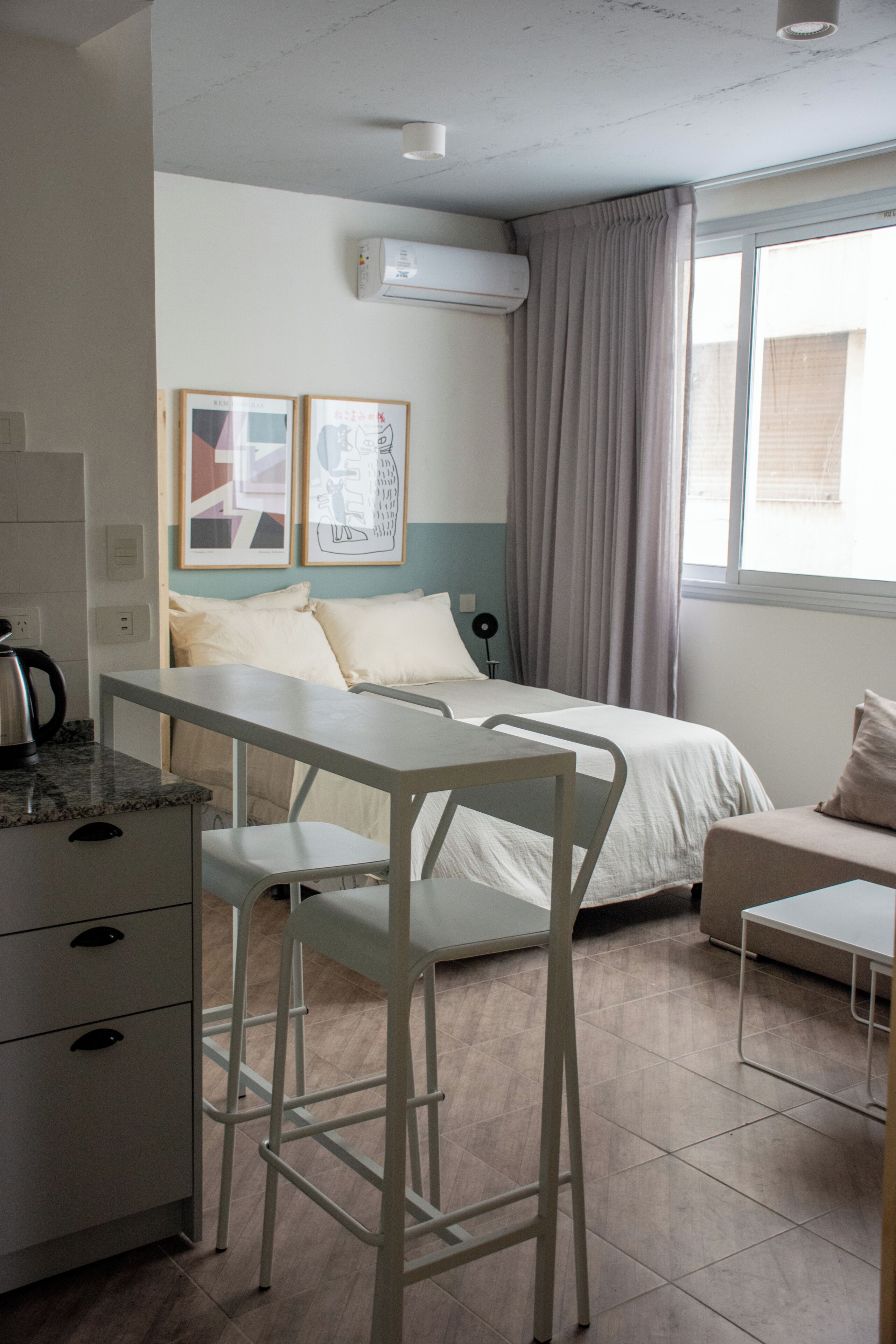 A modern, well-lit bedroom with a bed, a desk, and a chair. There is a window with curtains, a radiator, and artwork on the wall.