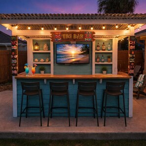 Un bar de style tiki avec un comptoir en bois, trois tabourets, un écran de télévision diffusant une image de coucher de soleil, et des étagères remplies de bouteilles et d’objets de décoration.