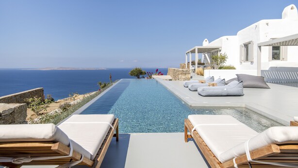 A poolside area with lounge chairs, a stone wall, and a view of the sea. - VILLA FAROS NEAR PORT, MYKONOS BY MYKONOS AGENDA (Mykonos)