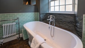 Une salle de bain avec une baignoire sur pieds, une fenêtre donnant sur un mur en pierre et un radiateur.