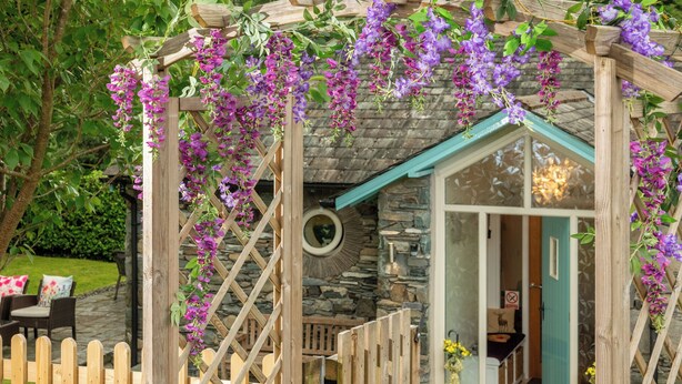 A wooden arbor with purple flowers, a stone wall, and a door leading to a room with a blue door and a mirror on the wall. - Overbeck, Ambleside - Two Bedroom Cottage, Sleeps 4 (Ambleside)