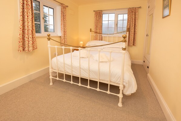 Une chambre avec un lit simple, un cadre en métal blanc, deux fenêtres avec des rideaux, un sol recouvert d’une moquette et une petite table de chevet avec une lampe.