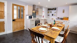 A dining area with a wooden table set for four, surrounded by chairs. There is a microwave and oven in the kitchen area to the left. - Juniper Grasmere - Juniper Cottage, Grasmere (Grasmere)