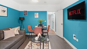 Una sala de estar moderna con una pared de acento color turquesa, un televisor de pantalla plana mostrando 'NETFLIX', un sofá gris, una mesa de centro y un comedor con sillas y una mesa.