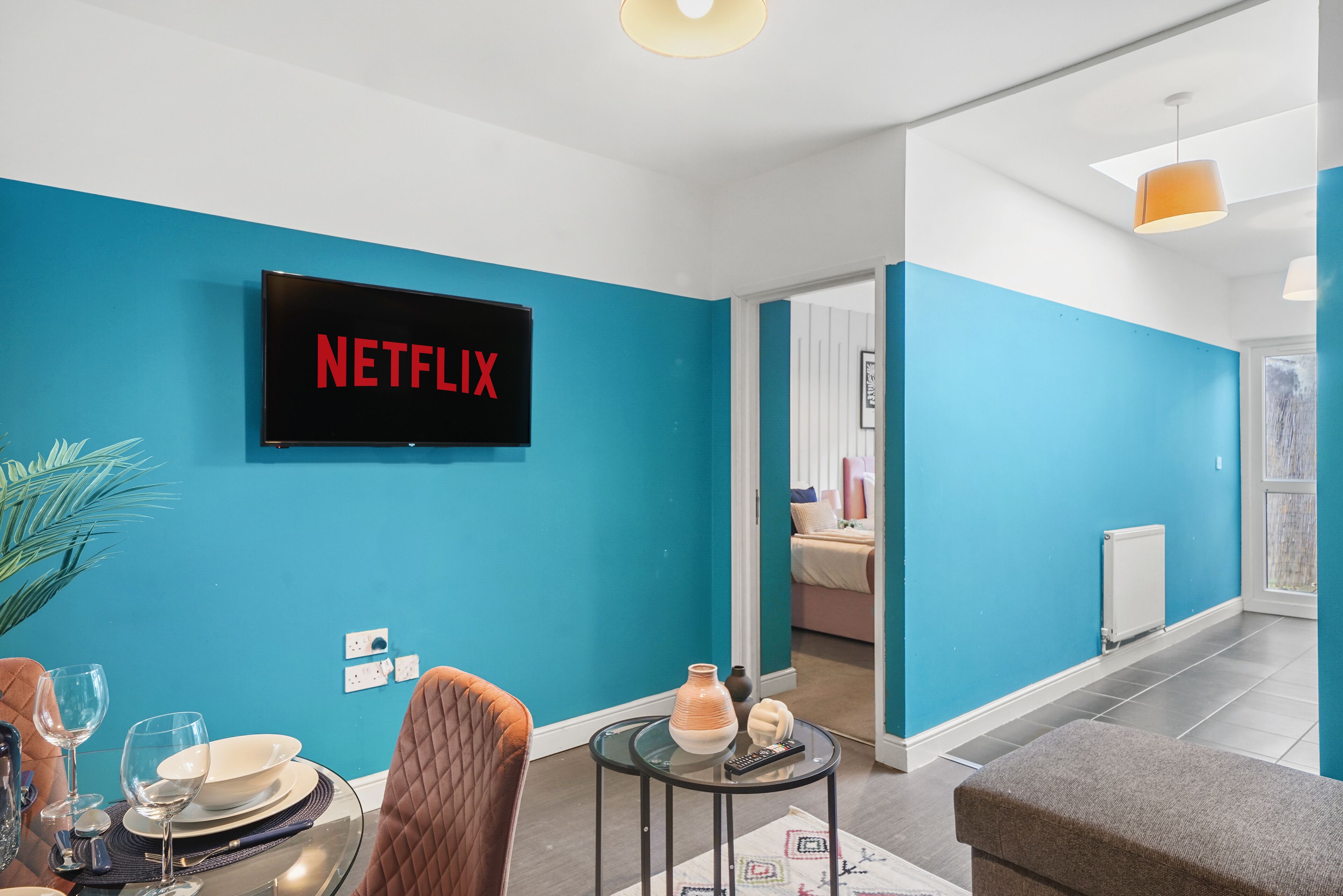 A modern living room with a teal wall, a flat-screen TV displaying "NETFLIX," a dining area with a table set for two, and a cozy seating area with a brown sofa.