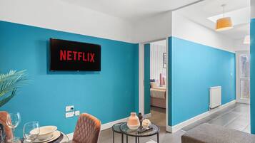 A modern living room with a teal wall, a flat-screen TV displaying "NETFLIX," a dining area with a table set for two, and a cozy seating area with a brown sofa.
