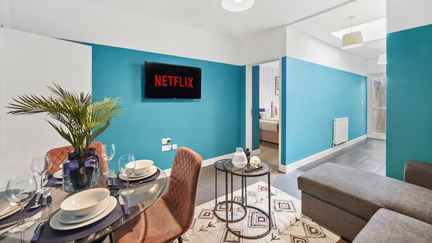 Una sala de estar moderna con una pared azul decorativa, un letrero de Netflix, un comedor con mesa de vidrio y una zona de estar acogedora.