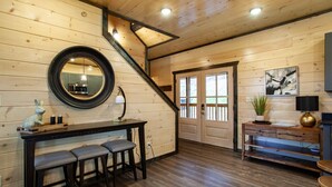 A wooden interior with a staircase, a round mirror, a dining area with stools, a small table with a lamp, and a door leading outside. - New 5BR Cabin w/ Pool, Hot Tub • Near Smokies (Sevierville)