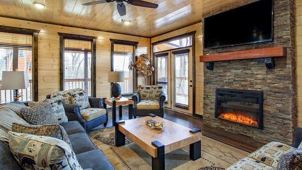 A cozy living room with a stone fireplace, wooden ceiling, and large windows. - New 5BR Cabin w/ Pool, Hot Tub • Near Smokies (Sevierville)