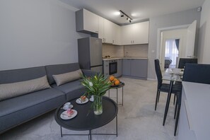 A modern living room with a grey sofa, a coffee table with a vase and fruit, and a kitchen in the background. - COOP Apartments (Sofia)