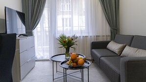 Un salón moderno con un sofá gris, una mesa de centro negra con fruta, un televisor en un mueble y cortinas traslúcidas.