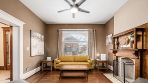 A living room with a yellow sofa, wooden flooring, a ceiling fan, a fireplace, and a window with curtains. - Stately Executive Home with Modern Amenities (Columbus)